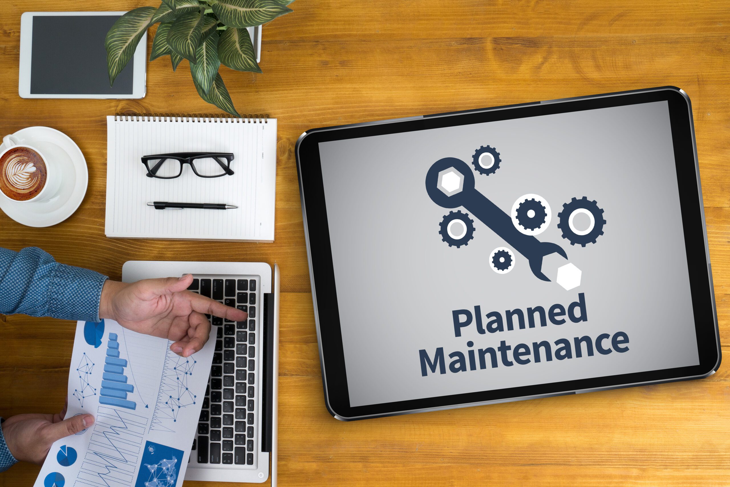 Aerial view planned maintenance text on a tablet next to laptop, pen and paper, latte