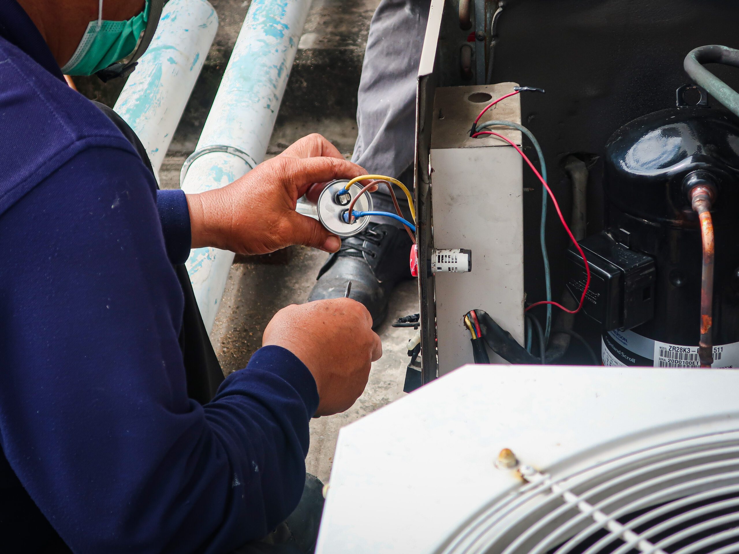 Technician performing HVAC repair, replacing a capacitor in an air conditioning unit with exposed wiring and compressor components.