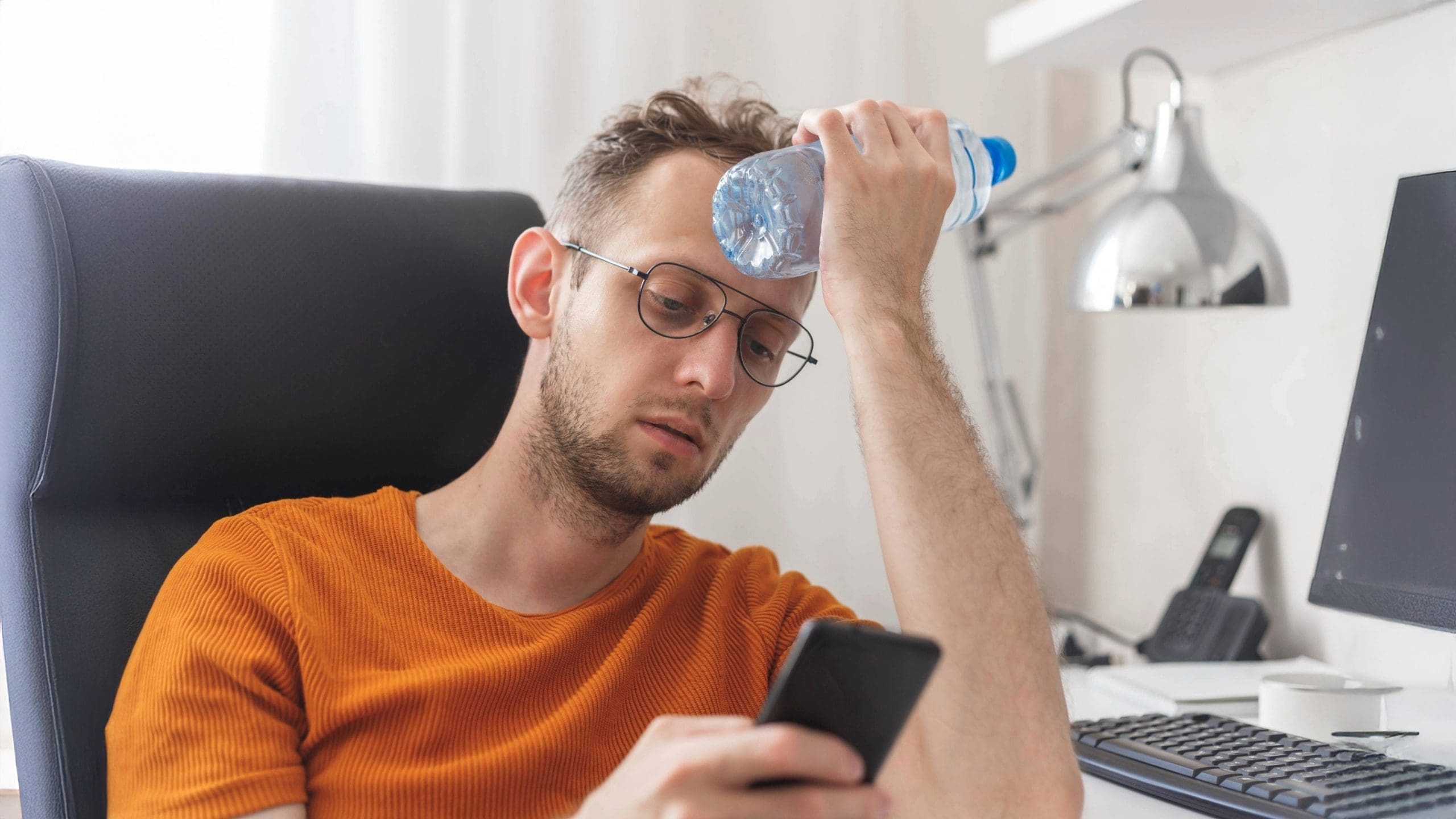 A man in an orange shirt sits uncomfortably at his desk, pressing a cold water bottle to his forehead while looking at his phone—likely searching for "heating and cooling near me" after his AC stopped working.