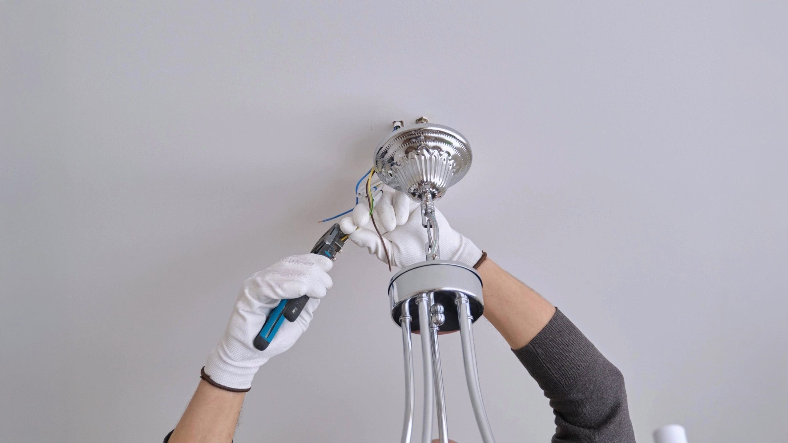 Electrician wearing white gloves installs a ceiling light fixture, using pliers to connect electrical wires on a white ceiling.